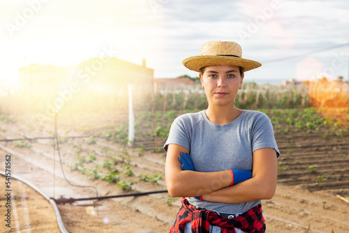 Smiling successful young female horticulturist posing against background of farm field with growing potatoes bushes and irrigation system on sunny summer day..