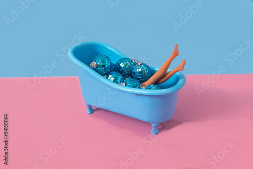 A blue bathroom tube full of blue shiny Christmas disco balls and women legs against pink and blue background. Surreal funny concept for end of the year festivities banner or card or print.  photo