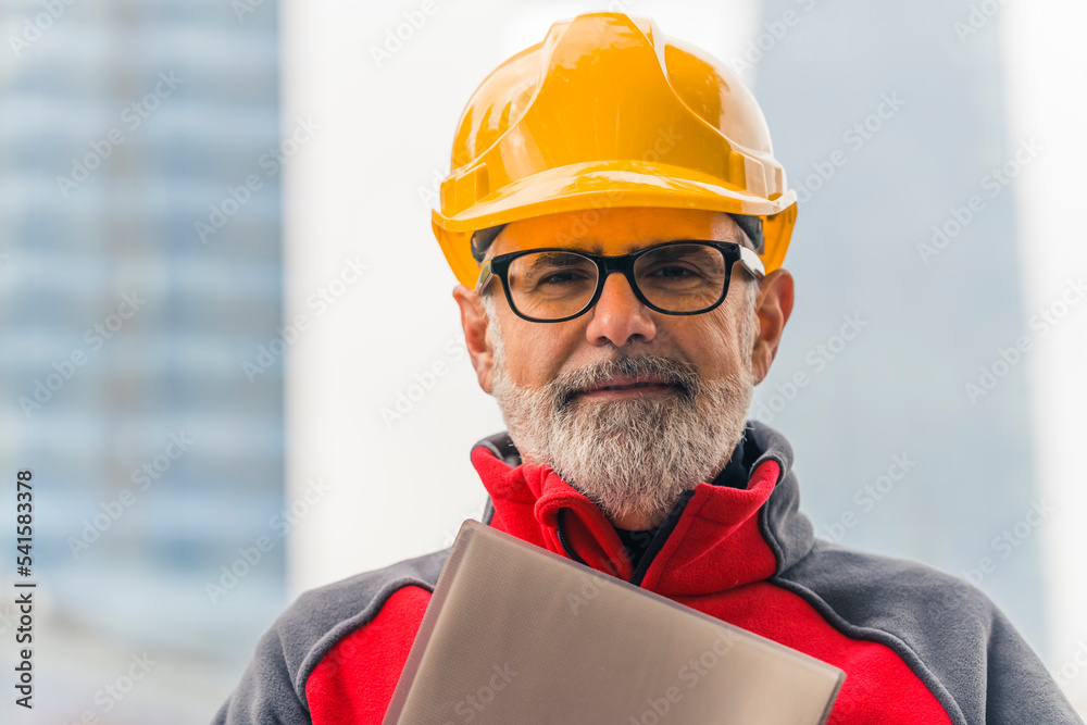Portrait of a confident, caucasian, bearded, grey-haired, middle-aged professional construction worker in uniform wearing a bright orange helmet and eyeglasses, holding a folder with documents Portrait of a confident, caucasian, bearded, grey-haired, middle-aged professional construction worker in uniform wearing a bright orange helmet and eyeglasses, holding a folder with documents