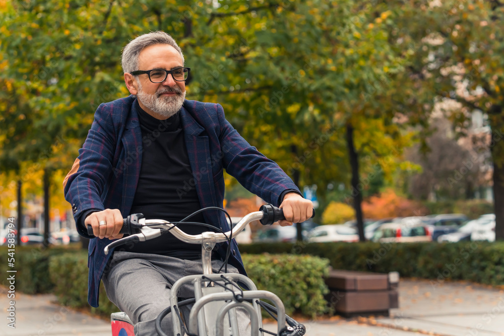 Middle-aged grey-haired man in smart-casual clothes briding bike to wokr through city with trees in background. Healthy lifestyle. Horizontal outdoor shot. High quality photo