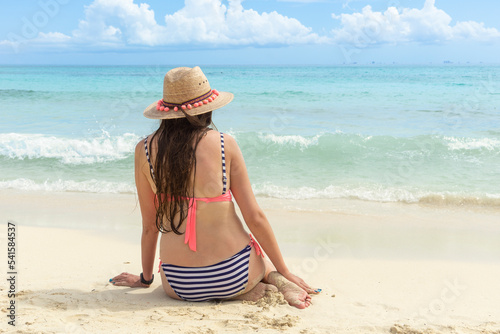 A girl in a bikini and with long hair sits on the beach with her back to the camera and looks at the sea. A girl in a hat and swimsuit looks at the waves and the seascape. The concept of loneliness.