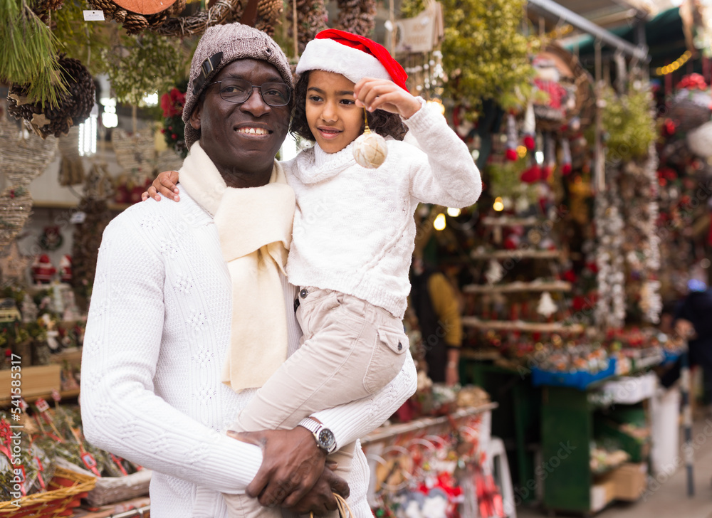 Portrait of joyful small girl with father in hat with Christmas toys at fair outdoor