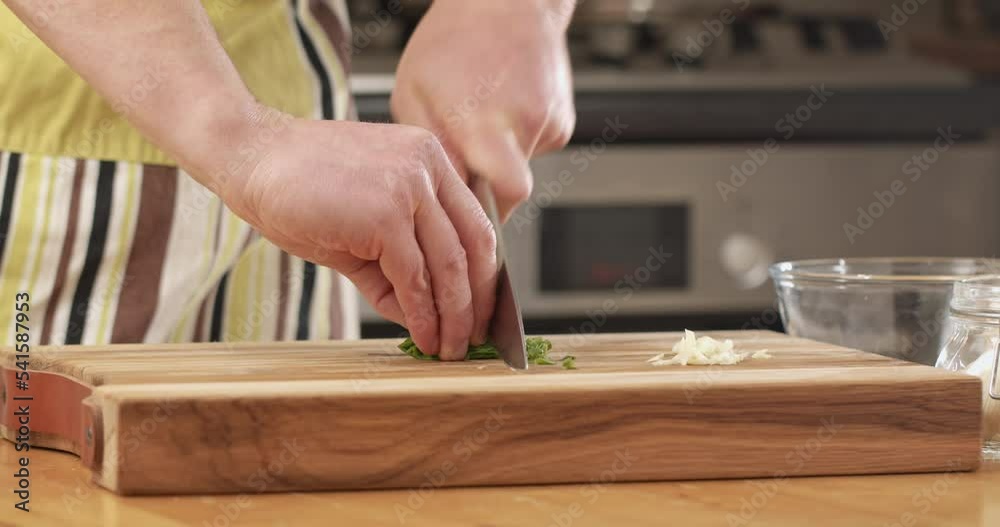 Male home cook chopping fresh basil leaves on a cutting board in his kitchen. Closeup shot. Male home cook chopping fresh basil leaves on a cutting board in his kitchen. Closeup shot.