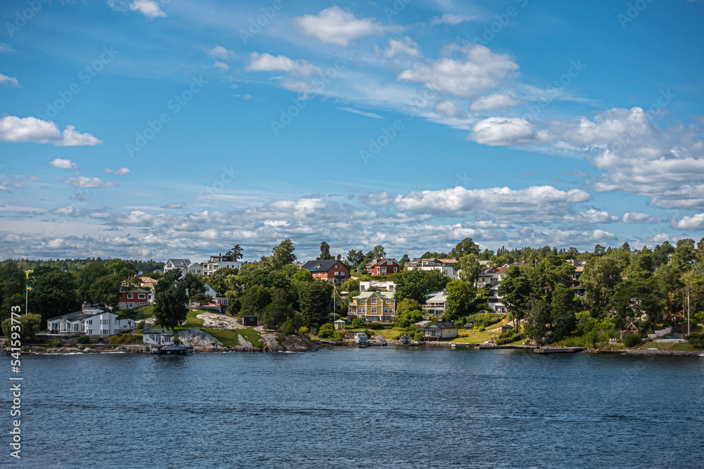 Sweden, Stockholm - July 17, 2022: Kappala on Lidingo island. Shoreline near Utkiksberget under blue cloudscape. Landscape hiding mansions between green and a few motor boats on water