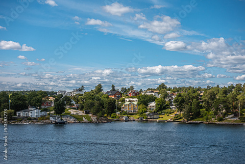 Sweden, Stockholm - July 17, 2022: Kappala on Lidingo island. Shoreline near Utkiksberget under blue cloudscape. Landscape hiding mansions between green and a few motor boats on water photo