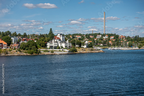 Sweden, Stockholm - July 17, 2022: Kappala on Lidingo island. Neptunivagen street reaches dock and Baltic water under blue cloudscape. Red roofs and Käppalaförbundet chimney, photo