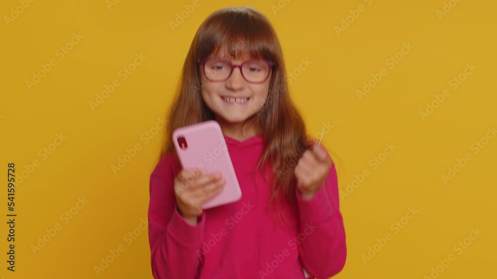 Young preteen child girl kid using credit bank card and smartphone while transferring money, purchases online shopping, order food delivery. Little toddler children alone on studio yellow background Young preteen child girl kid using credit bank card and smartphone while transferring money, purchases online shopping, order food delivery. Little toddler children alone on studio yellow background