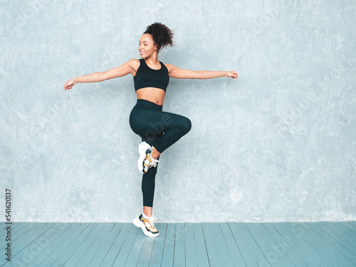 Portrait of fitness smiling black woman in sports clothing with afro curls hairstyle.Wearing sportswear.Sexy young beautiful model doing lunges before training.Female sitting in studio near grey wall