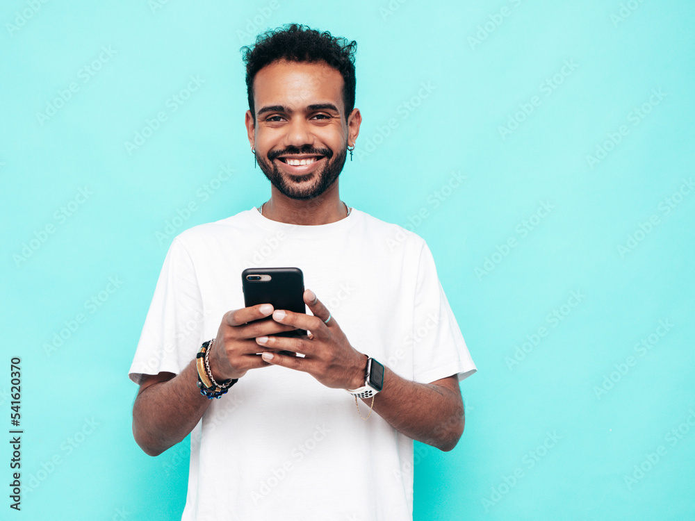 Handsome smiling hipster model. Sexy unshaven man dressed in summer t-shirt and jeans clothes. Fashion male posing near blue wall. Holding smartphone. Looking at cellphone screen. Using apps Handsome smiling hipster model. Sexy unshaven man dressed in summer t-shirt and jeans clothes. Fashion male posing near blue wall. Holding smartphone. Looking at cellphone screen. Using apps