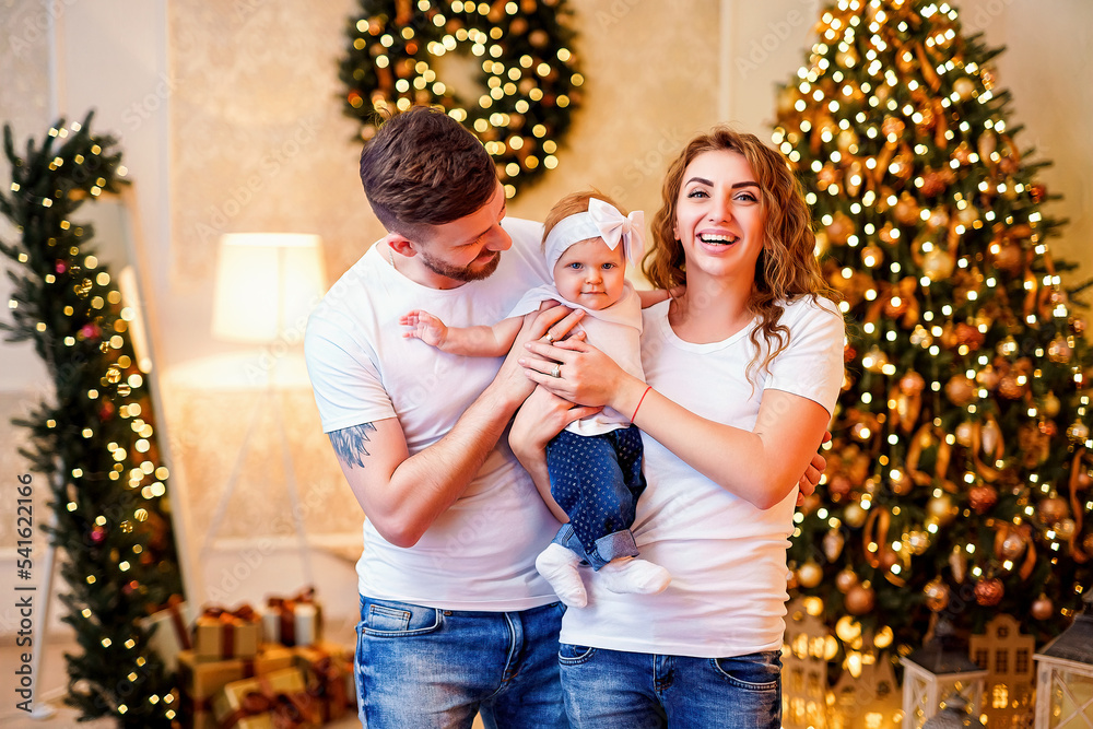 Happy young parents standing near Christmas tree in living room while holding little cute baby girl Happy young parents standing near Christmas tree in living room while holding little cute baby girl