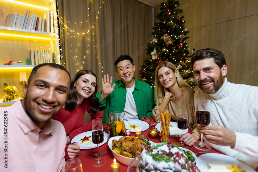 A company of interracial young people sitting at a festive New Year's table and taking a selfie together on a mobile phone. They hold glasses in their hands, wave at the camera, smile, rejoice. A company of interracial young people sitting at a festive New Year's table and taking a selfie together on a mobile phone. They hold glasses in their hands, wave at the camera, smile, rejoice.