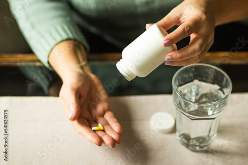 Elderly woman holding white plastic bottle and two capsules in hand, ready to take supplement pills with glass of water.