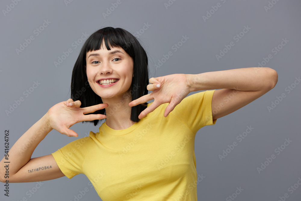 a happy, sweet woman stands smiling at the camera on a gray background in a yellow T-shirt and shows a victory sign with her fingers holding her hands near her face a happy, sweet woman stands smiling at the camera on a gray background in a yellow T-shirt and shows a victory sign with her fingers holding her hands near her face