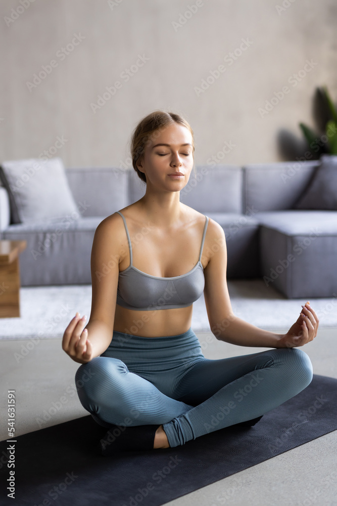 Young healthywoman in sportive top and leggings practicing yoga at home sitting in lotus pose on yoga mat meditating smiling relaxed with closed eyes, Mindfulness meditation concept Young healthywoman in sportive top and leggings practicing yoga at home sitting in lotus pose on yoga mat meditating smiling relaxed with closed eyes, Mindfulness meditation concept