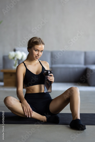 Beautiful fit woman drinks a glass of water at home after a workout inside her apartment.