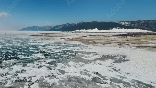 Drone moves over the frozen lake towards the mountains. Snow and cracked ice form a unique pattern. A clear blue sky merges on the horizon with the surface of the ice. The first spring days in Baikal