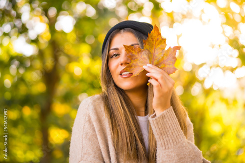 Young blonde girl with a wool cap in autumn  with a leaf on her face at sunset in a natural park