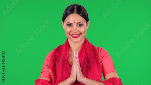 Portrait of a young Indian girl with red bindi dot in national classic red sari looking straight, smiles and folded palm to palm. A brunette with long hair and red lips on green screen at studio. photo