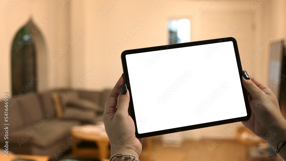 Hands holding tablet with blurred living room lighted with soft lights in background. Blank screen for your advertise design Hands holding tablet with blurred living room lighted with soft lights in background. Blank screen for your advertise design