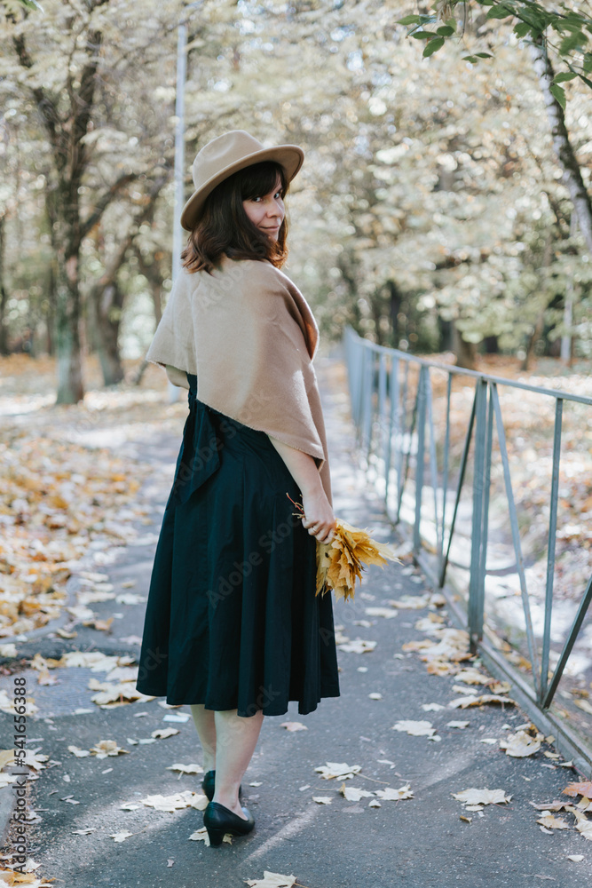 middle-aged woman in autumn felt hat and a bouquet of maple leaves walk in the park in the fall season, brown autumn tones