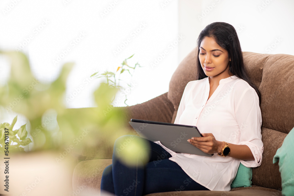 Pretty Indian young woman using digital tablet sitting on sofa at home. Pretty Indian young woman using digital tablet sitting on sofa at home.