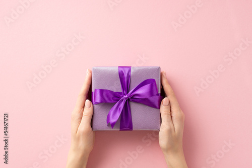 New Year concept. First person top view photo of woman's hands giving lilac giftbox with purple ribbon bow on isolated pastel pink background