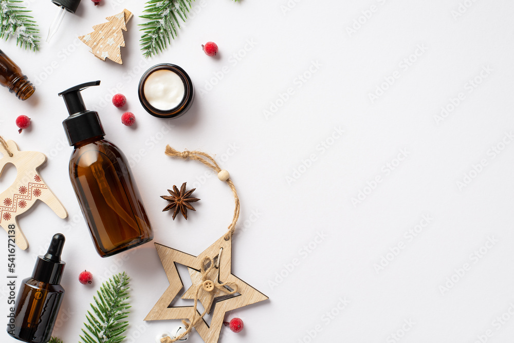 Winter skincare products concept. Top view photo of amber glass bottles cream jar christmas wooden ornaments fir branches and mistletoe berries on isolated white background with copyspace Winter skincare products concept. Top view photo of amber glass bottles cream jar christmas wooden ornaments fir branches and mistletoe berries on isolated white background with copyspace