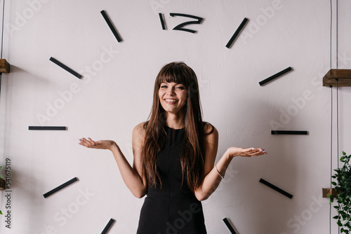 Playful brunette young woman in black dress standing at home against wall with big clock spreads hands in hesitate, toothy smiling looking at camera. Hispanic woman using hands like a clock hands. photo