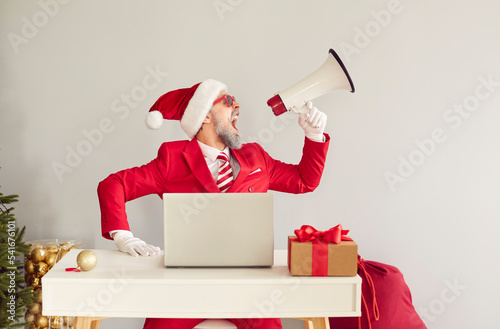 Funny excited man wearing red suit, Santa Claus cap and sunglasses sitting at office desk with laptop and gift box, advertising online Christmas holiday sale and yelling loudly through megaphone photo