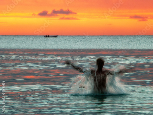 Silhouette of a man splashing in Indian Ocean at sunset with a boat in distance, Mauritius