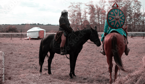 Two warriors are on horseback. Warrior in a helmet, armor and with a shield. Against the background of a green field and a white yurt. The concept of the Middle Ages, battles