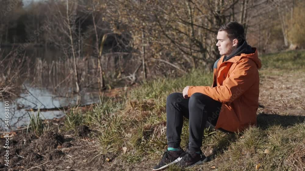 Young man, with an orange jacket, sits near the lake and contemplates the panorama Young man, with an orange jacket, sits near the lake and contemplates the panorama