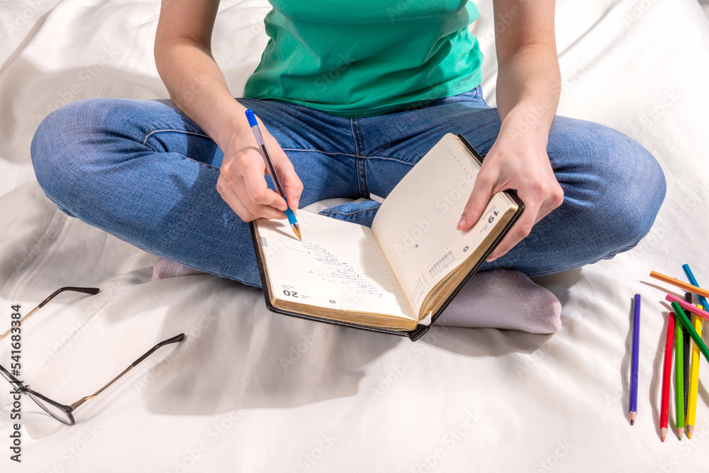 Obraz premium A girl in a green T - shirt and blue jeans is sitting on the bed and writing in a notebook , colored pencils and glasses are scattered around