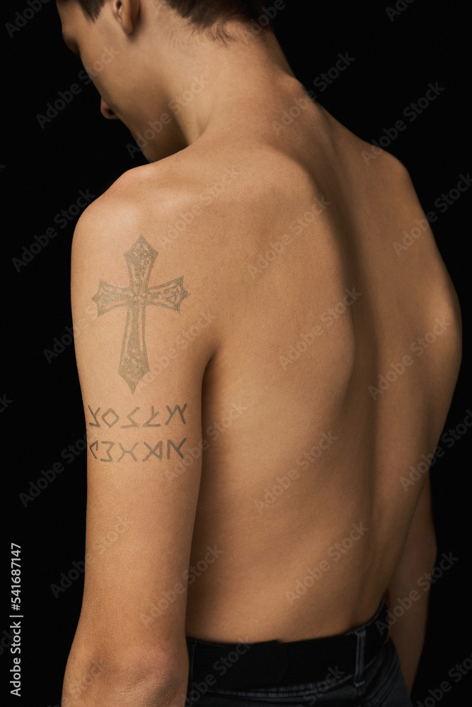 Naklejka premium Close-up shot of a young man with a water-based temporary cross tattoo on his shoulder. The portrait of a shirtless tattooed young man standing on a dark background. Back view.