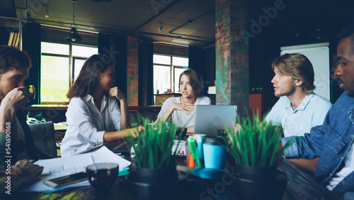 Fotografia Young woman is speaking with colleagues sitting at table in modern office discussing work plans while her team is listening to her, male coworker is drinking tea