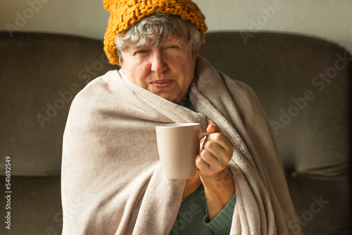 Elderly woman in ginger cap wrapped in blanket and drinking cup of tea. Old grandma feeling cold at home. Energy crisis.