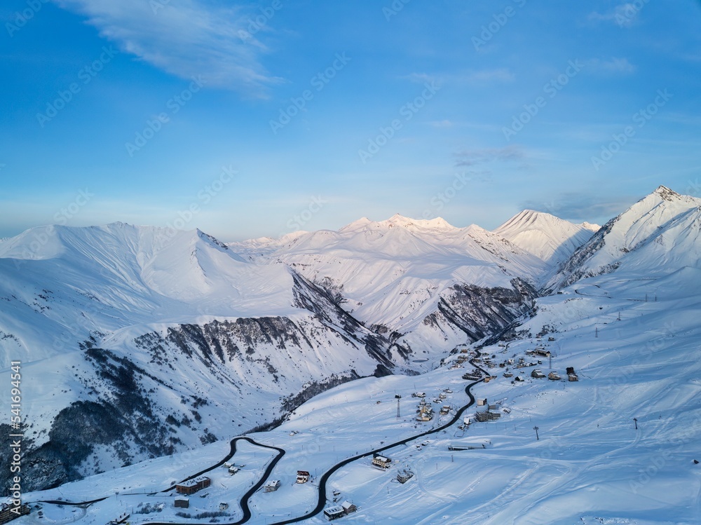 Obraz premium Wide aerial panorama of snowy mountain ridge at ski resort village on sunrise. Stunning mountains range covered with snow powder on ski resort at sunset. Caucasus mountain peaks skyline on sunset.