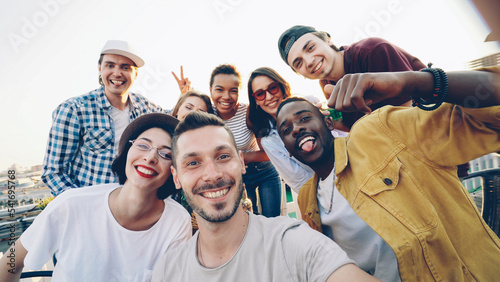 Point of view shot of bearded young man looking at camera taking selfie with friends sitting on roof at table and having fun.