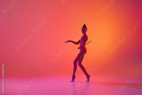 Studio shot of young charming girl  rhythmic gymnast training with sports equipment isolated over pink background in neon light filter