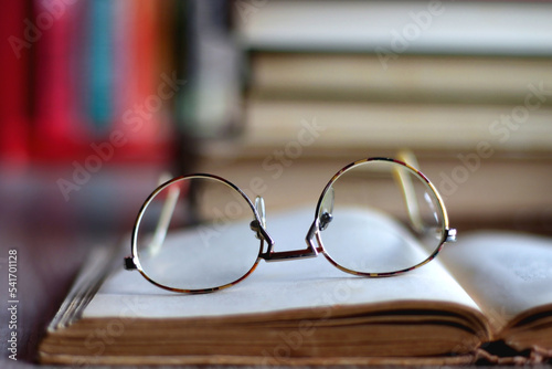 Stack of colorful hardcover books and reading glasses in front of a bookshelf. Selective focus.