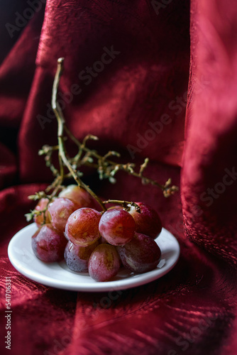 Bunch of grapes on a white plate against background of red luxury fabric. Vertical photo with grapes on the background of blurred red textile