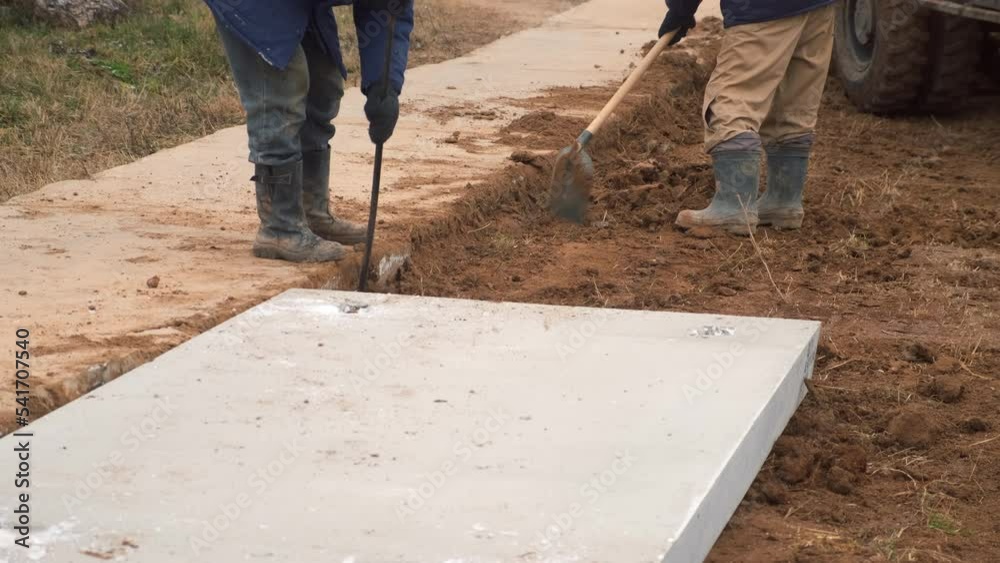 Two man construction workers with shovels dig the ground to lay cement concrete blocks, repair roads on the construction site. Legs of two builders in boots on the ground. Manual labour.