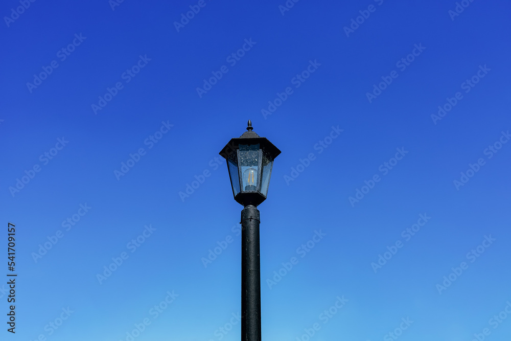 Dirty black lamppost with an electric bulb against a bright blue daytime sky. Horizontal background with sky and vintage city electric lantern in close-up