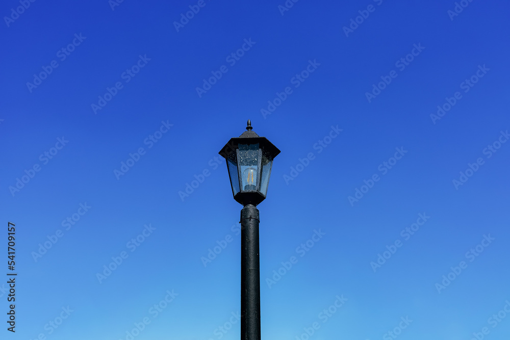 Dirty black lamppost with an electric bulb against a bright blue daytime sky. Horizontal background with sky and vintage city electric lantern in close-up Dirty black lamppost with an electric bulb against a bright blue daytime sky. Horizontal background with sky and vintage city electric lantern in close-up