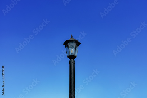 Dirty black lamppost with an electric bulb against a bright blue daytime sky. Horizontal background with sky and vintage city electric lantern in close-up