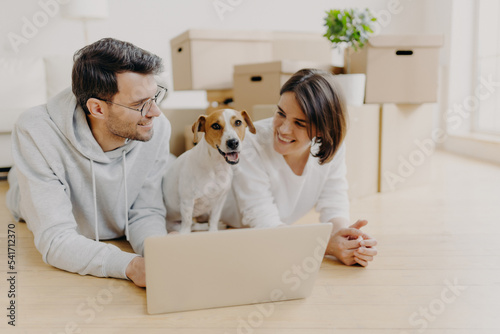 Lovely just married couple lie on floor, use laptop computer, creats design project for new house, their favourite dog poses between them, pile of cardboard boxes in background. Moving to new home