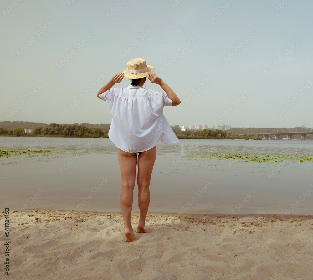 Rear view of a young woman in a black swimsuit and white shirt walking along the beach near the water. A woman is resting on the sand in summer. Woman in a beach hat