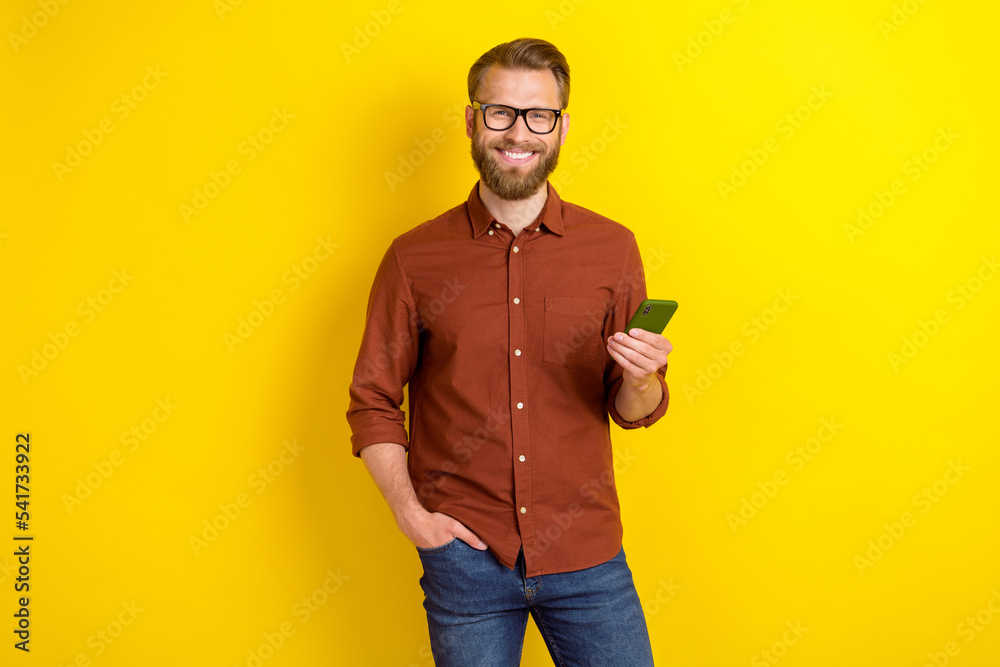 Portrait of cheerful funny glad man with masculine beard burgundy shirt hold smartphone arm in pocket isolated on yellow color background Portrait of cheerful funny glad man with masculine beard burgundy shirt hold smartphone arm in pocket isolated on yellow color background