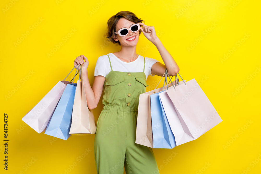 Photo of young cool celebrity woman wear khaki stylish overalls hold new sunglass many bags shopaholic smile isolated on yellow color background - obrazy, fototapety, plakaty 