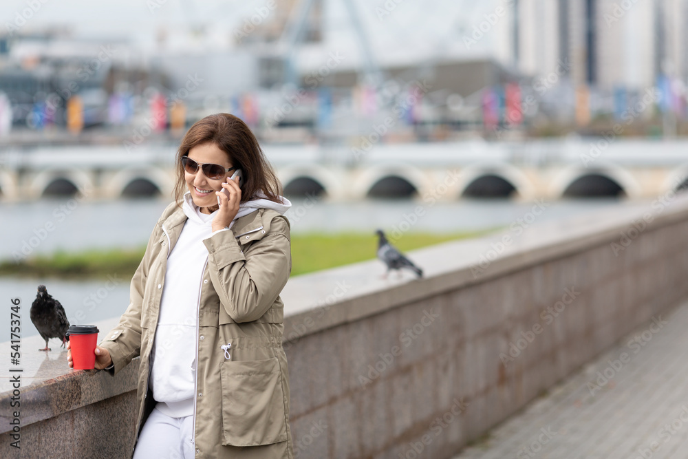 Fototapeta premium Attractive woman wearing dark glasses stands on the embankment talking on a mobile with coffee in hand and pigeons are nearby, selective focus.